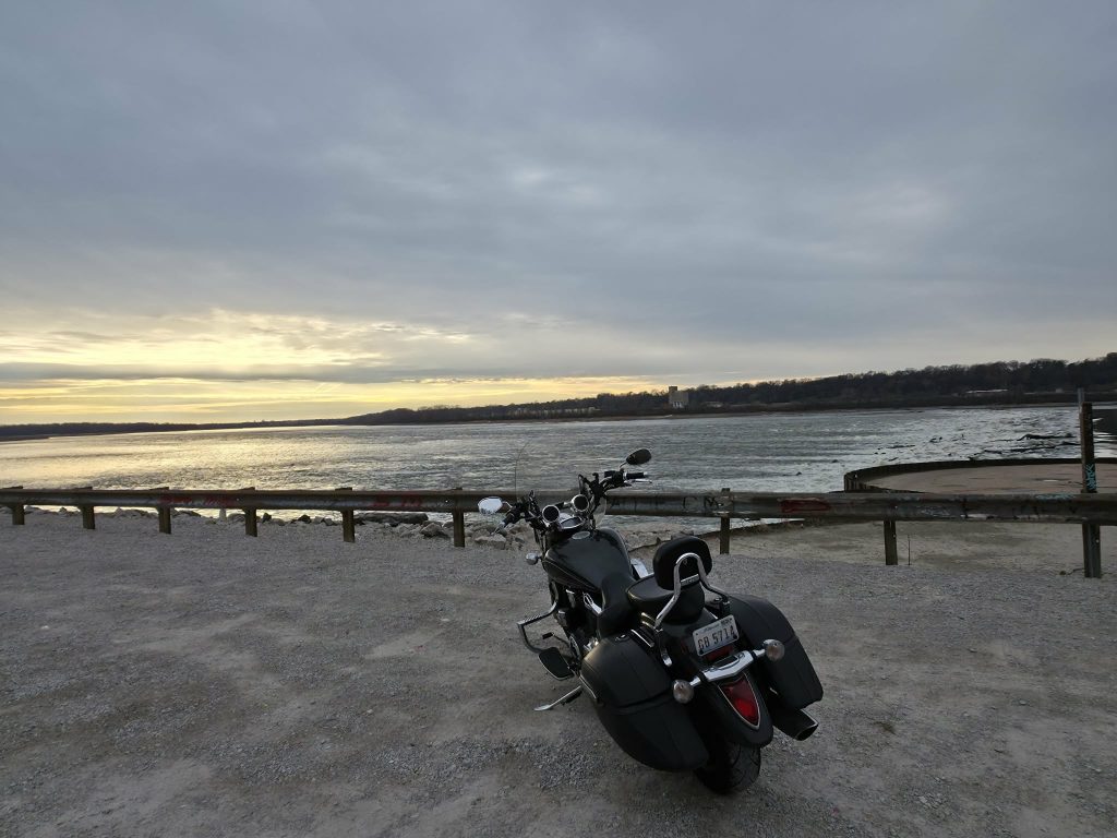 Black touring motorcycle parked on a gravel pull-off beside a wide river, gray overcast sky, bare trees on the far shore.