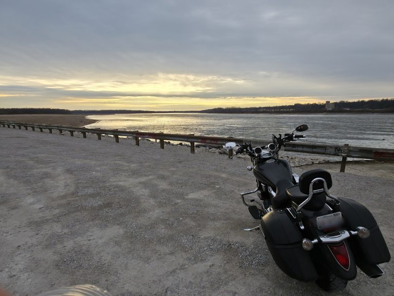 Black cruiser motorcycle parked by a wide river under an overcast sky, gravel bank and bare far shore