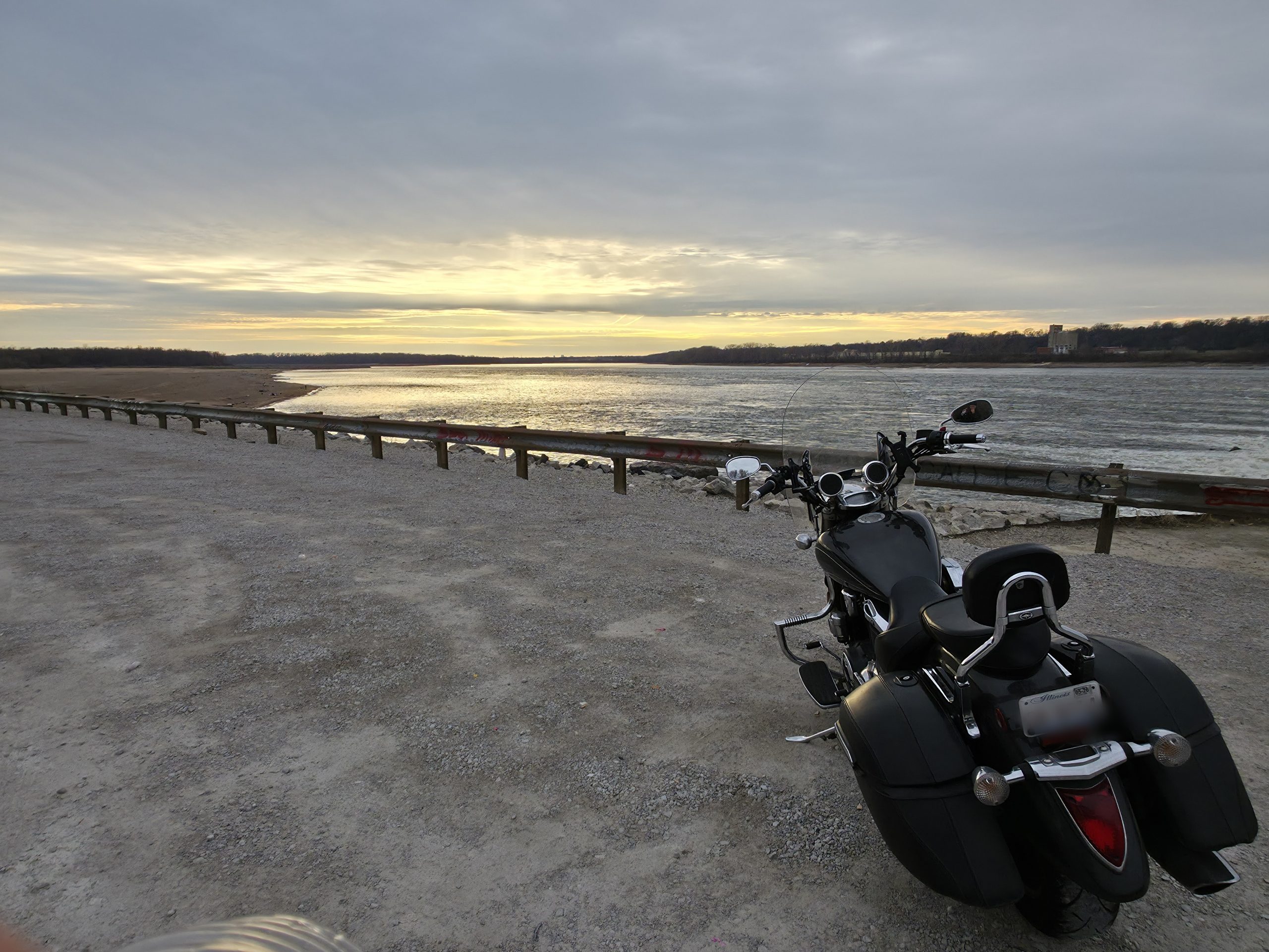 Black cruiser motorcycle parked by a wide river under an overcast sky, gravel bank and bare far shore