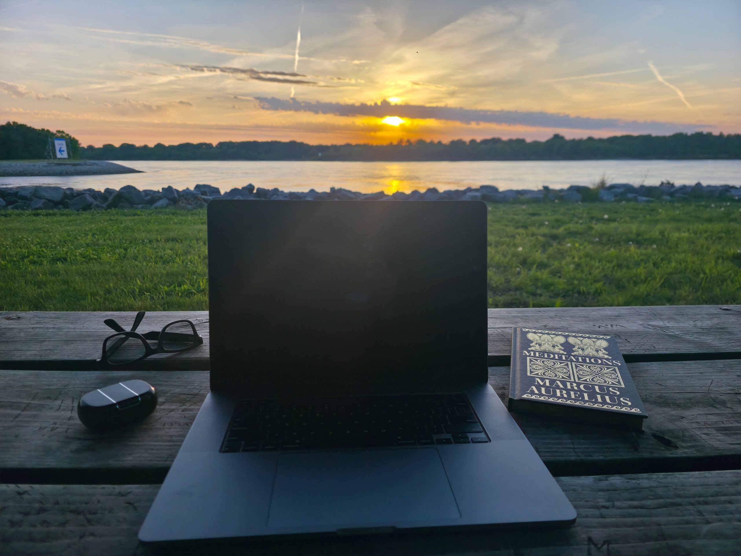 Open laptop and a worn copy of Marcus Aurelius's Meditations on a picnic table beside a calm lake at sunset