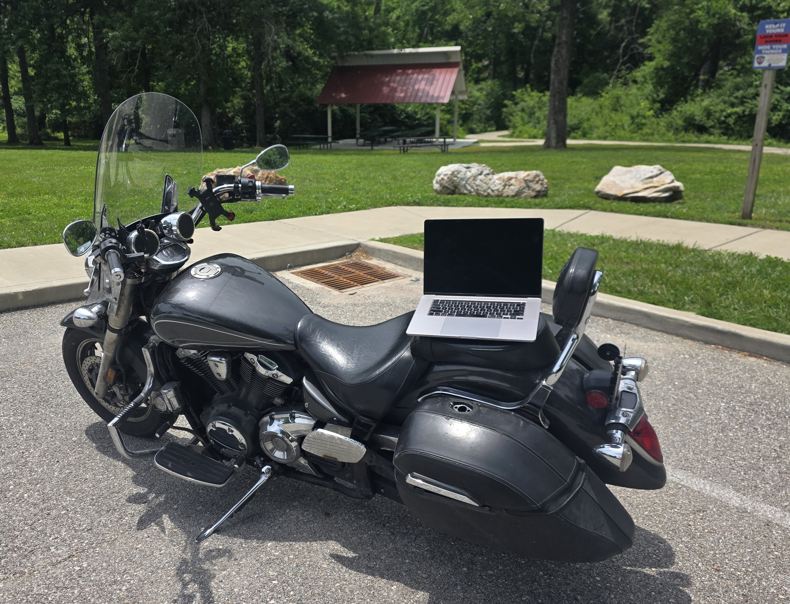 Open laptop resting on the seat of a parked cruiser motorcycle in a quiet park, trees and a shelter in the background