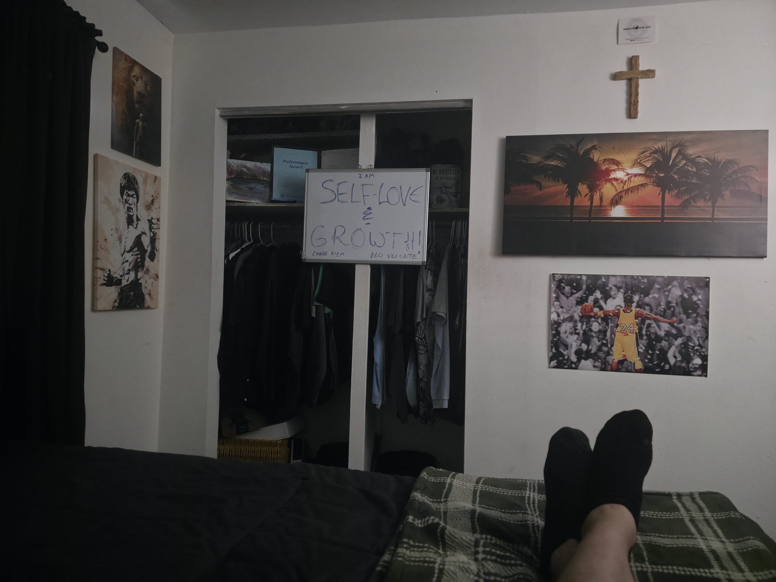 Dimly lit bedroom wall — cross, Bruce Lee, Kobe, and a handwritten 'Self-Love & Growth' whiteboard, viewed from the bed