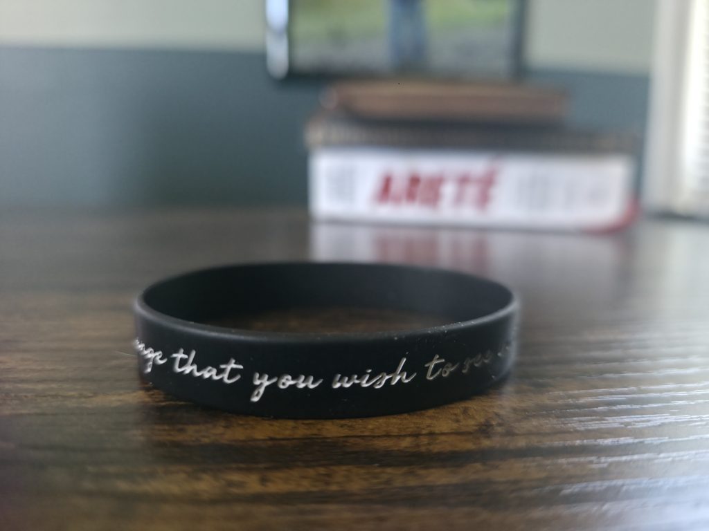 The same worn bracelet on the desk, this angle showing the fragment "be the change that y—" before the lettering fades.