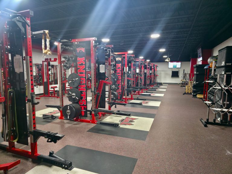 Empty high school weight room with rows of red power racks lit by shafts of overhead light