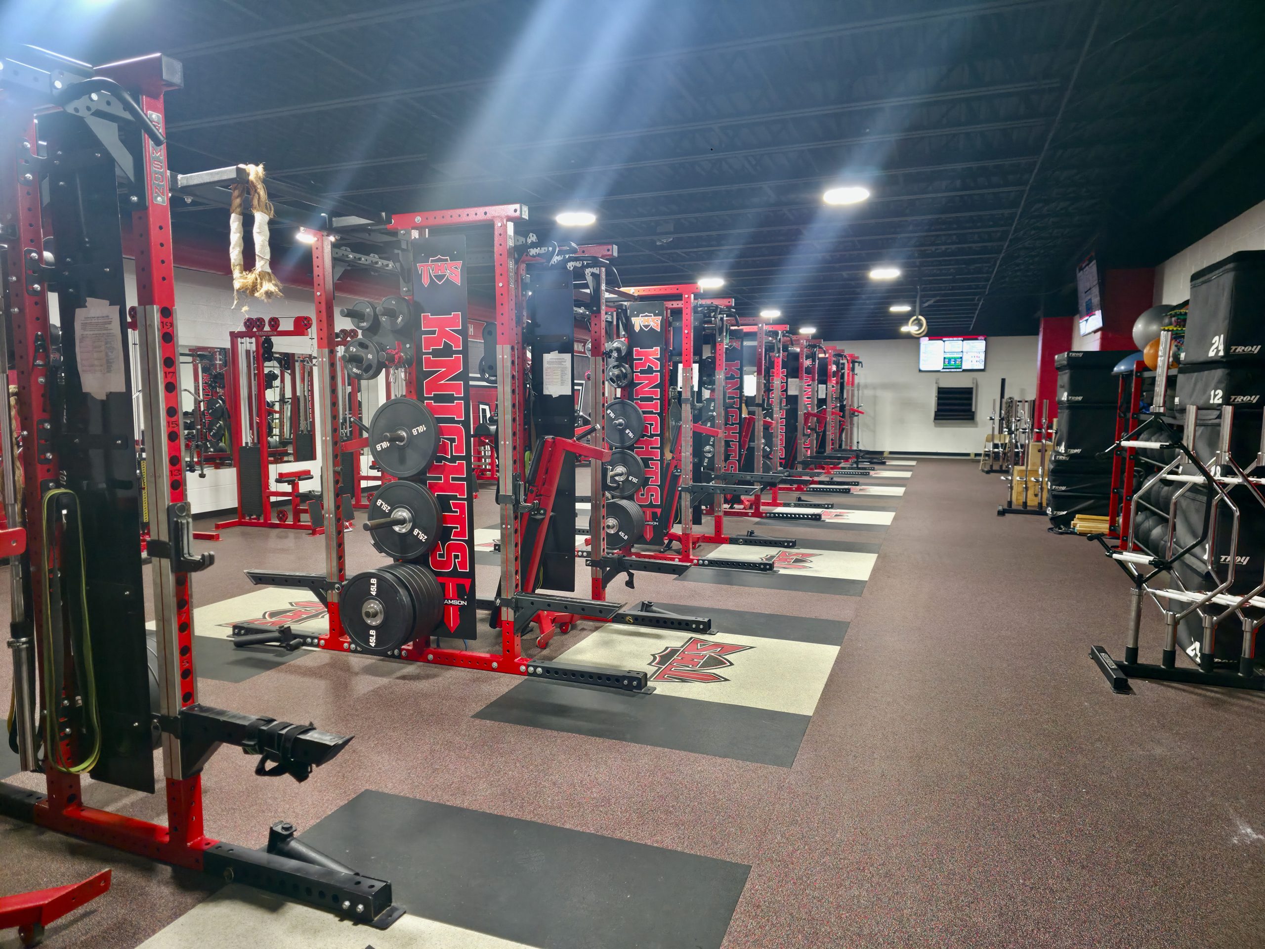 Empty high school weight room with rows of red power racks lit by shafts of overhead light