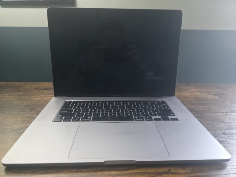 Open six-year-old MacBook laptop on a wooden desk against a plain wall, screen off in soft natural light