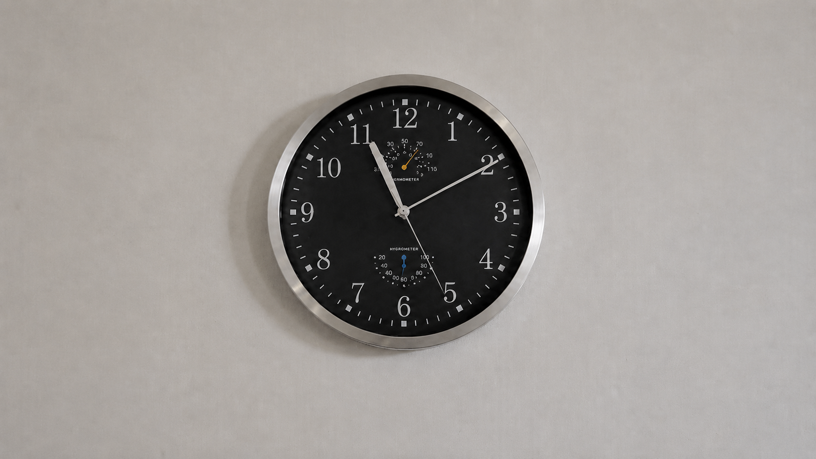Round wall clock with a black face and brushed-metal rim, small thermometer and hygrometer dials inside