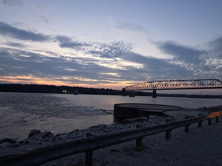 Steel truss bridge over the Mississippi River at dusk, churning current along a rocky, guardrail-lined bank