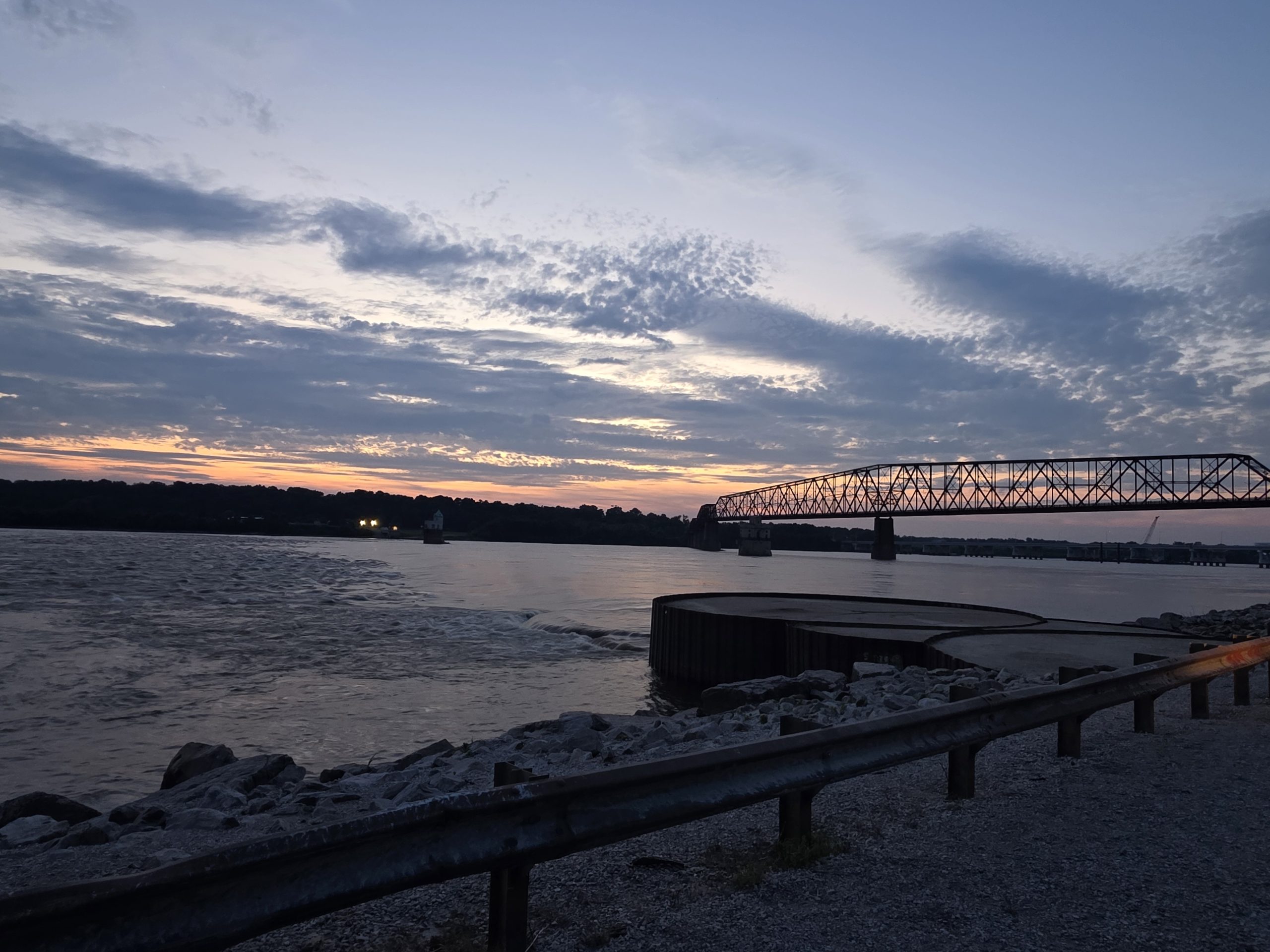 Steel truss bridge over the Mississippi River at dusk, churning current along a rocky, guardrail-lined bank