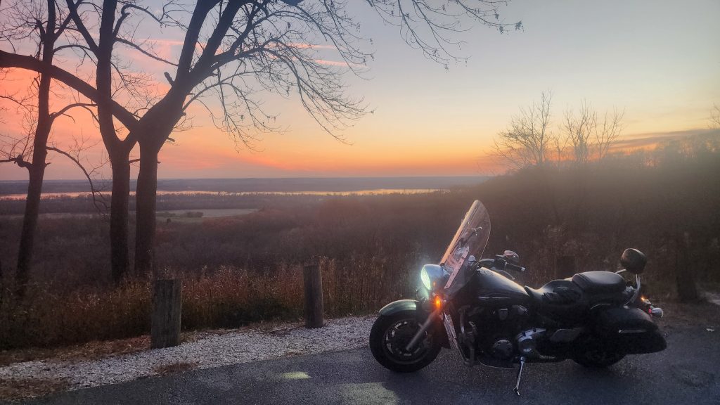 Motorcycle parked at a scenic overlook at dusk, pastel sky over the river valley below