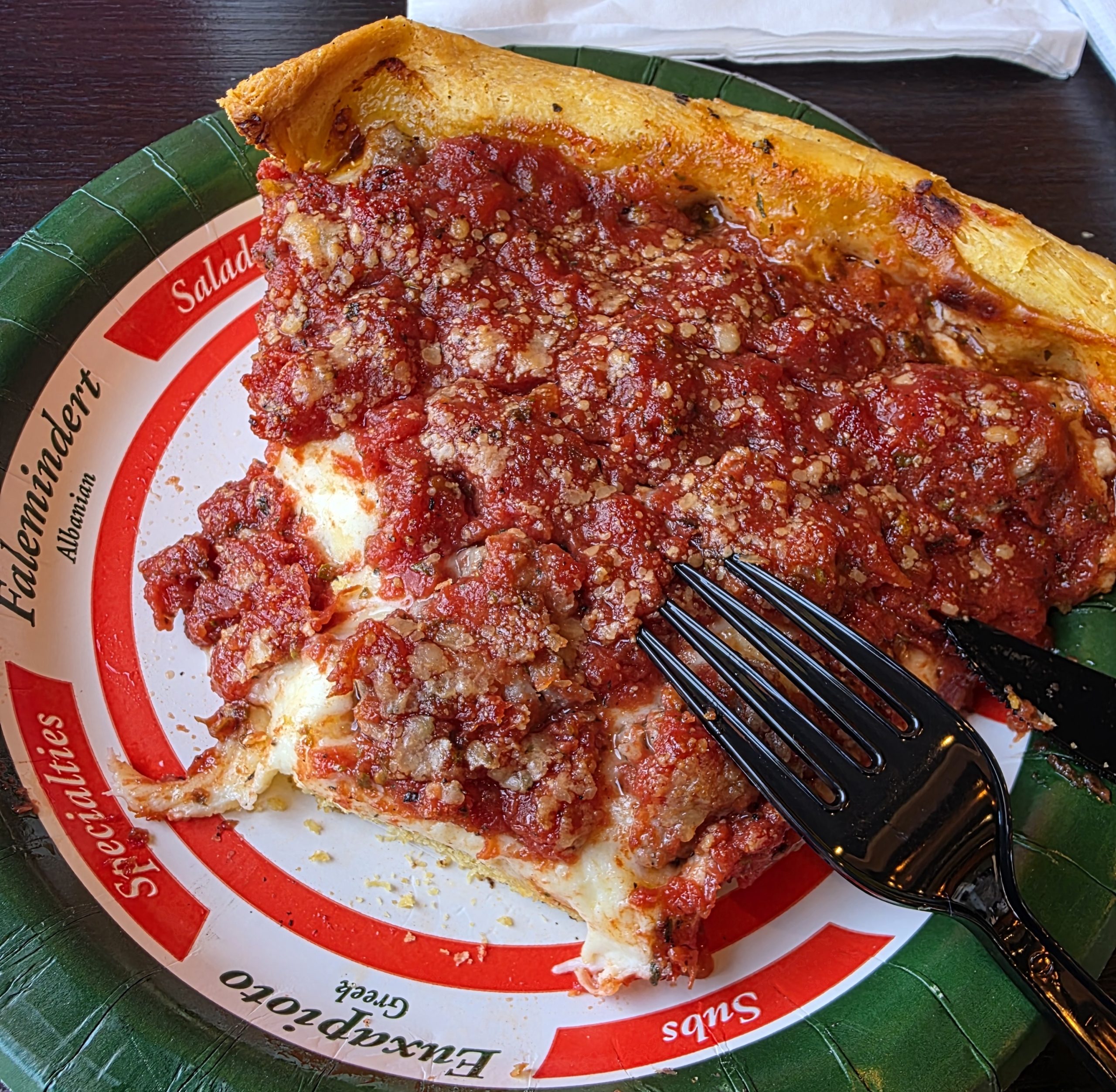 A thick slice of Chicago deep dish pizza with sausage and chunky tomato sauce on a branded pizzeria plate, fork resting on the side