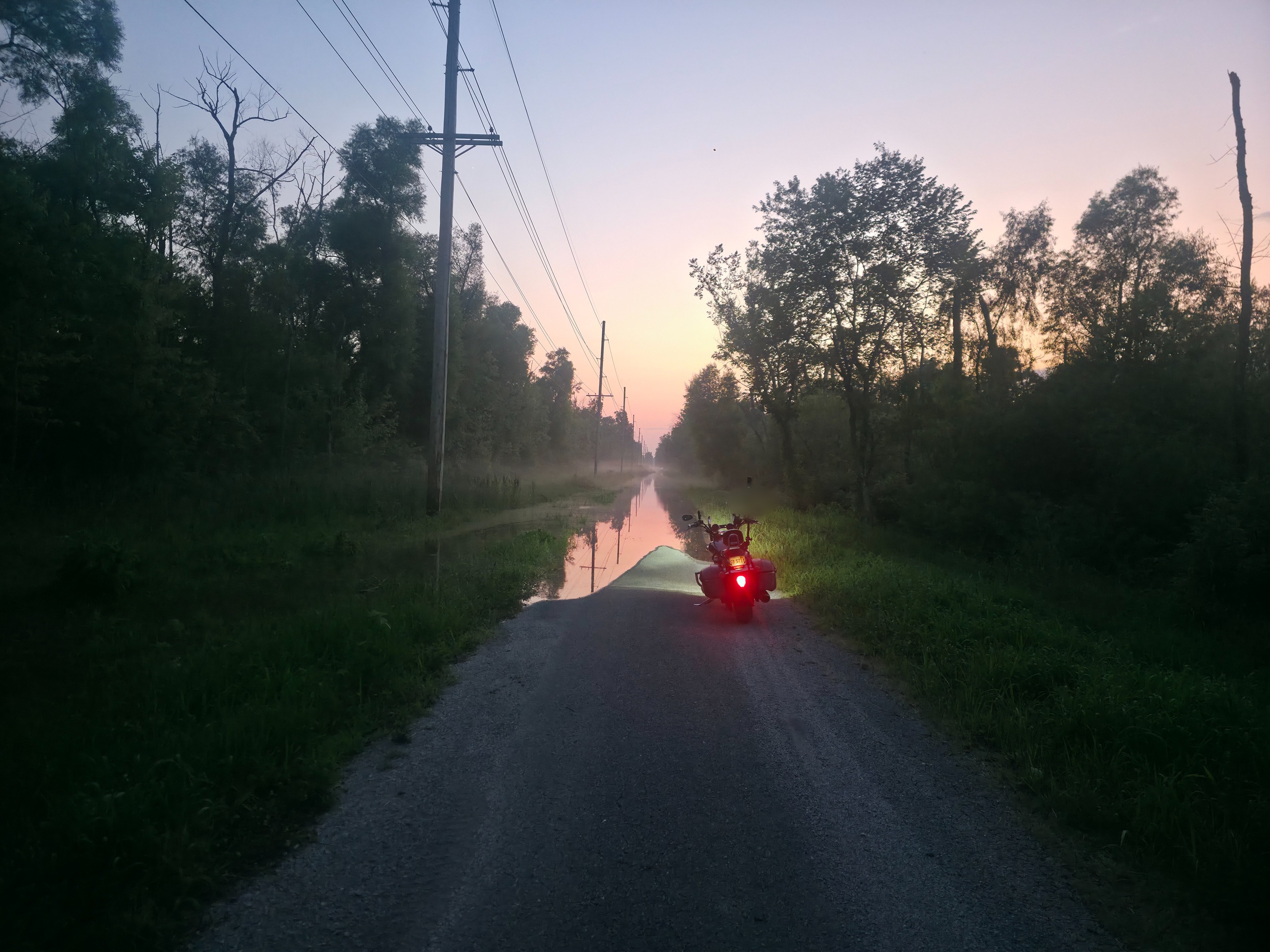 Motorcycle heading down a misty rural road at dusk, tail-light glowing, soft pink and orange light on the horizon
