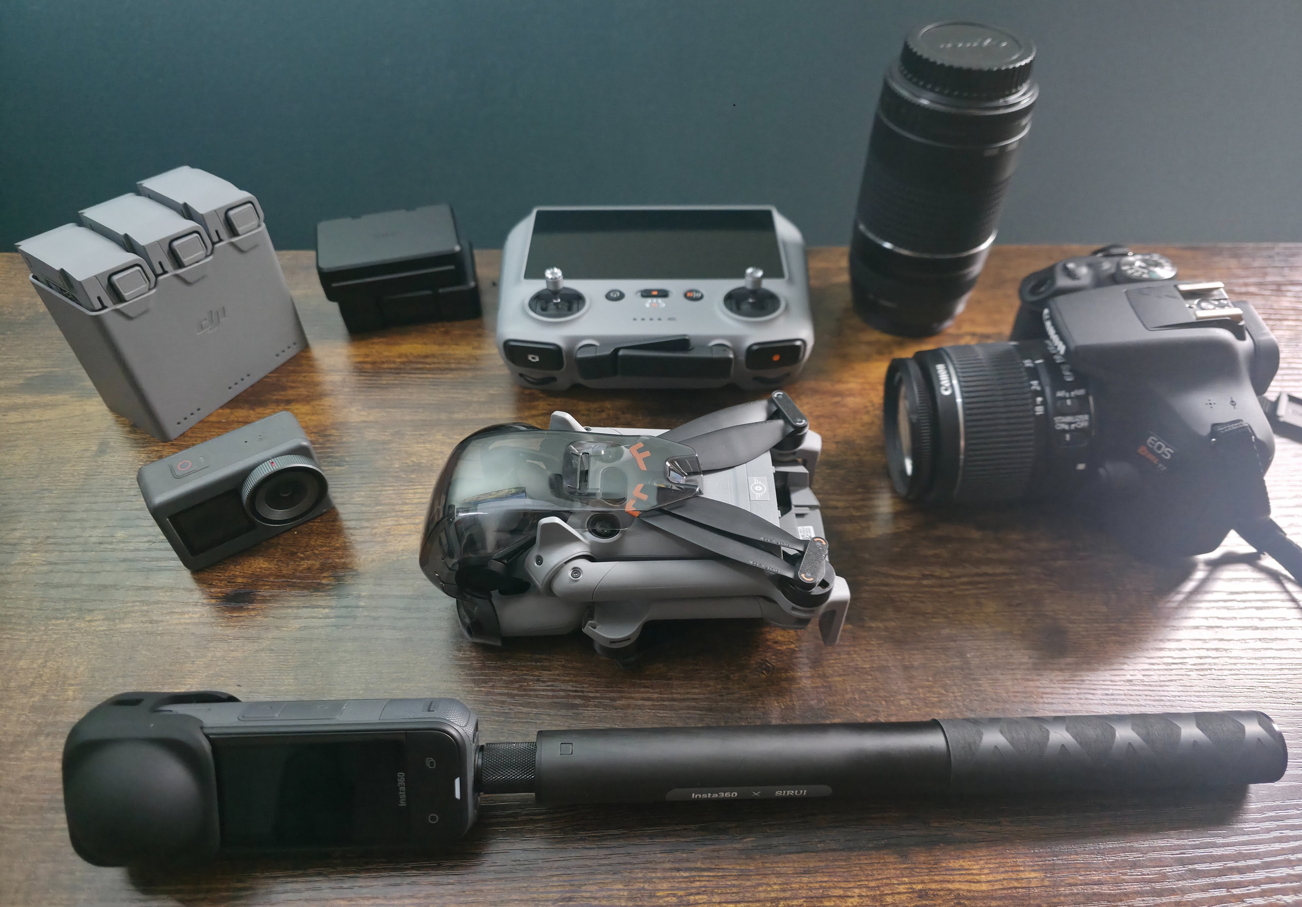 Camera gear laid out on a wooden desk: a DSLR with kit lens, a telephoto lens, a folded drone and controller, spare batteries, an action camera, and a 360 camera on a monopod