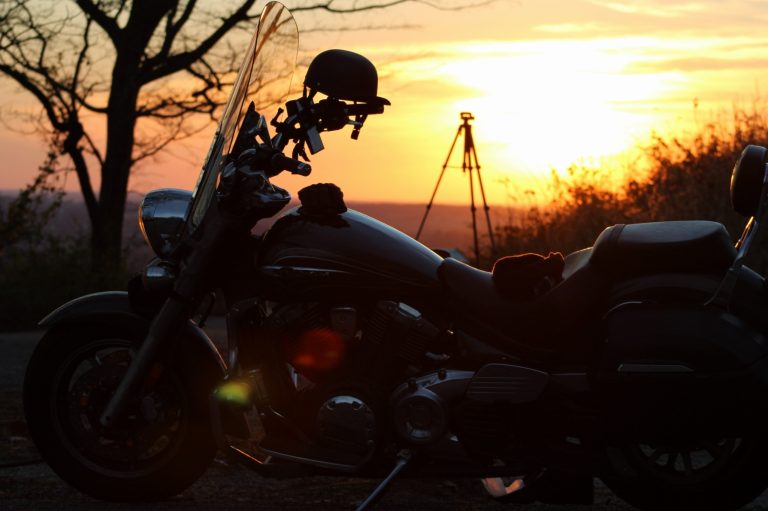 Motorcycle silhouetted against a golden sunset at a river overlook, helmet resting on the handlebars and a camera tripod in the background
