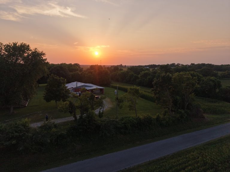 Aerial view of a red barn and gravel drive on a rural property at sunset, corn fields and treeline beyond, a figure standing near the road, taken on the drone's maiden flight