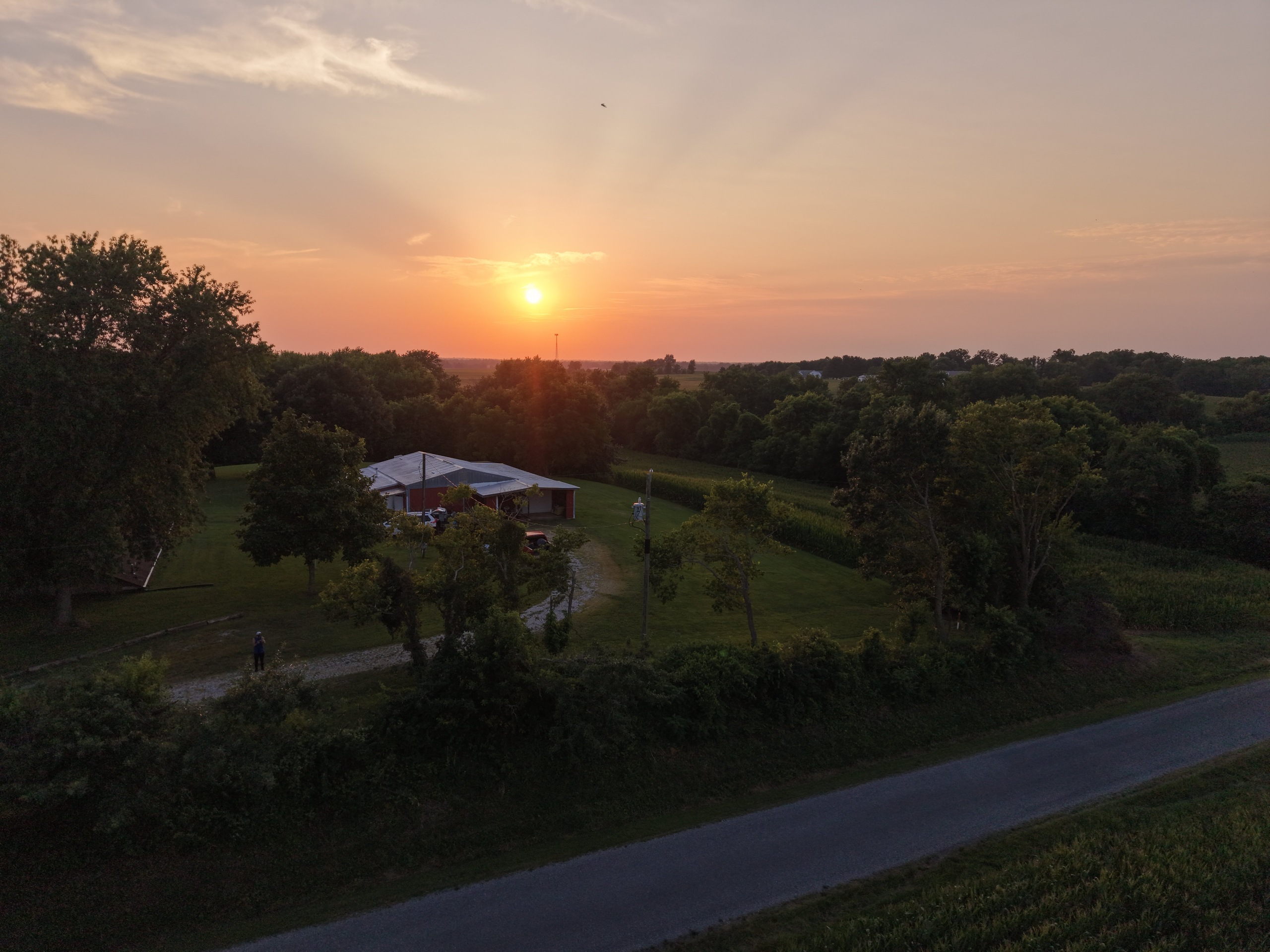 Aerial view of a red barn and gravel drive on a rural property at sunset, corn fields and treeline beyond, a figure standing near the road, taken on the drone's maiden flight