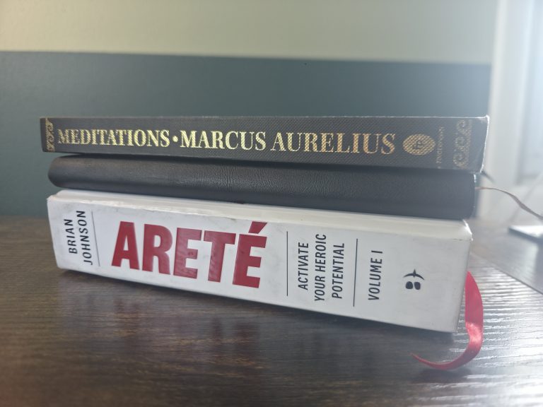 Marcus Aurelius's Meditations stacked with a worn copy of Brian Johnson's Areté on a wooden desk in low evening light.