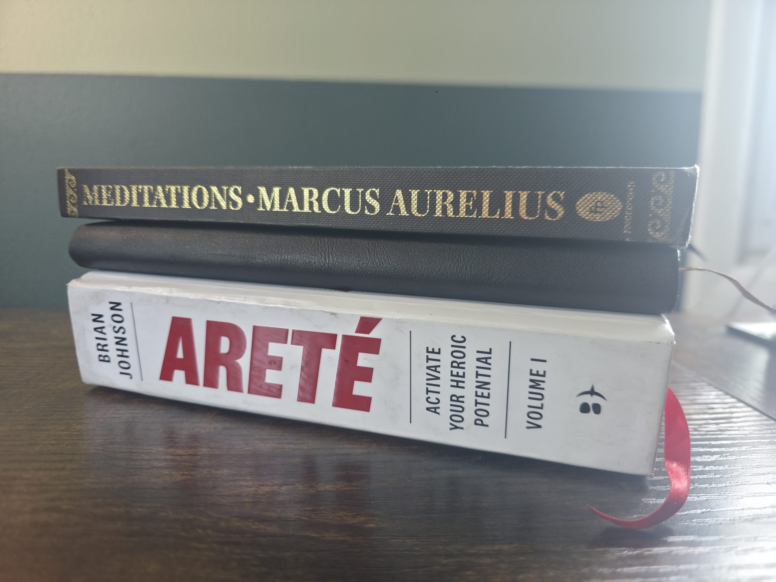 Marcus Aurelius's Meditations stacked with a worn copy of Brian Johnson's Areté on a wooden desk in low evening light.