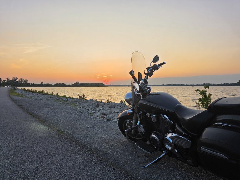 A black cruiser motorcycle parked at the water's edge at sunset, aimed down the shoreline road toward the horizon