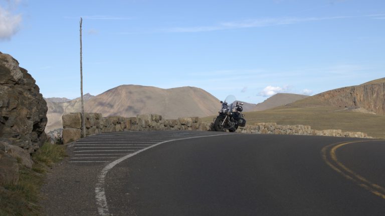 A lone touring motorcycle parked against a low stone wall on Trail Ridge Road, empty pavement curving through the foreground, bare snow-free tundra ridges behind under a pale blue sky.