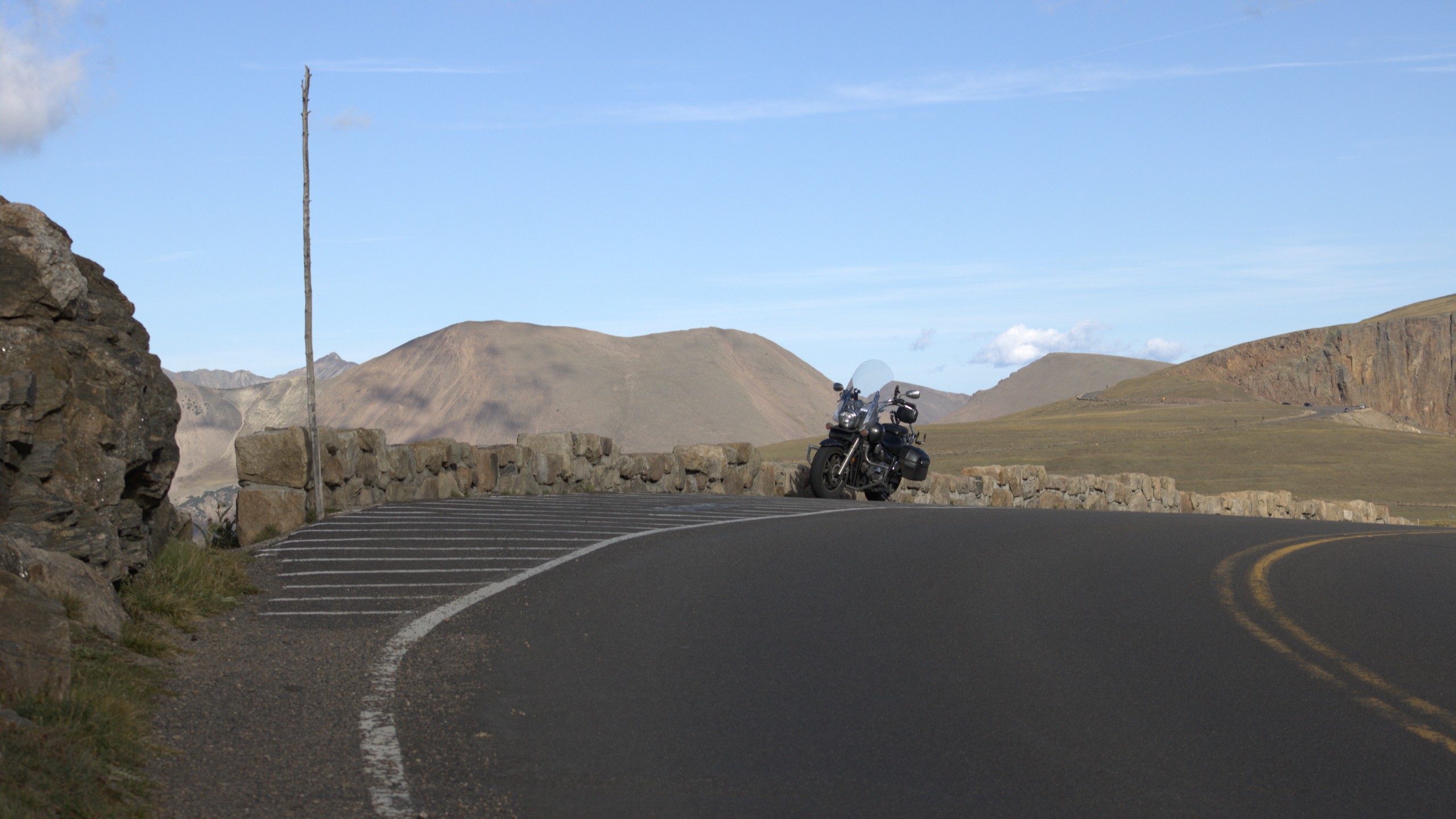 A lone touring motorcycle parked against a low stone wall on Trail Ridge Road, empty pavement curving through the foreground, bare snow-free tundra ridges behind under a pale blue sky.