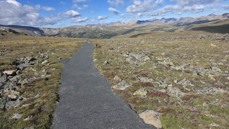 A narrow paved path running straight through open alpine tundra toward distant mountain ridges under a partly cloudy sky.