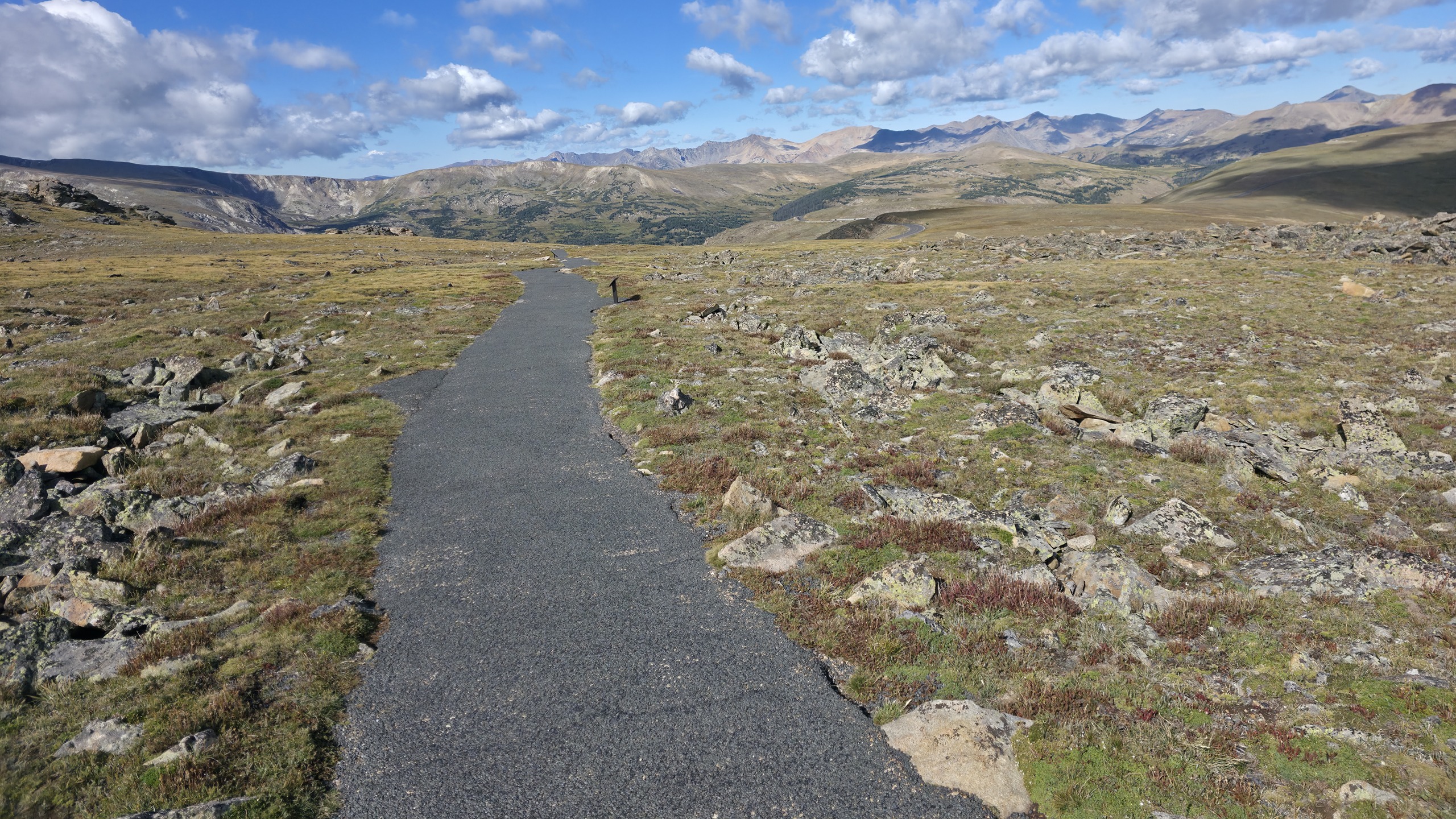 A narrow paved path running straight through open alpine tundra toward distant mountain ridges under a partly cloudy sky.