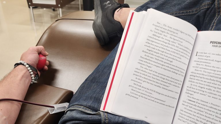 A man in a plasma donation chair with a paperback open across his lap, one hand squeezing a red ball, the tubing running along the armrest.
