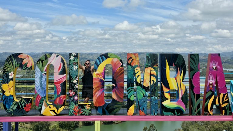 A man with both arms raised inside the giant painted COLOMBIA sign letters, green hills and the Guatapé reservoir behind them.